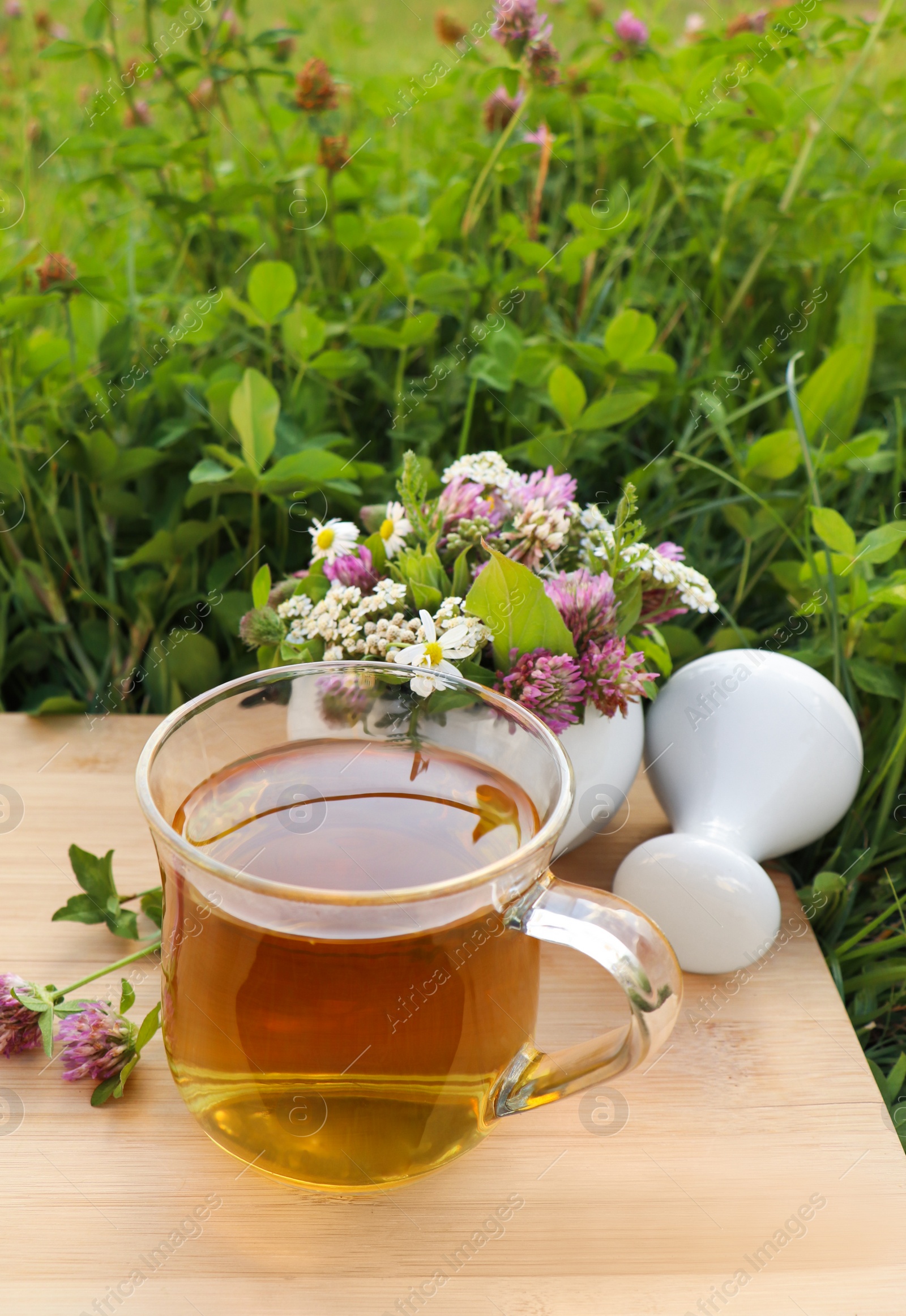 Photo of Cup of aromatic herbal tea, pestle and ceramic mortar with different wildflowers on wooden board in meadow