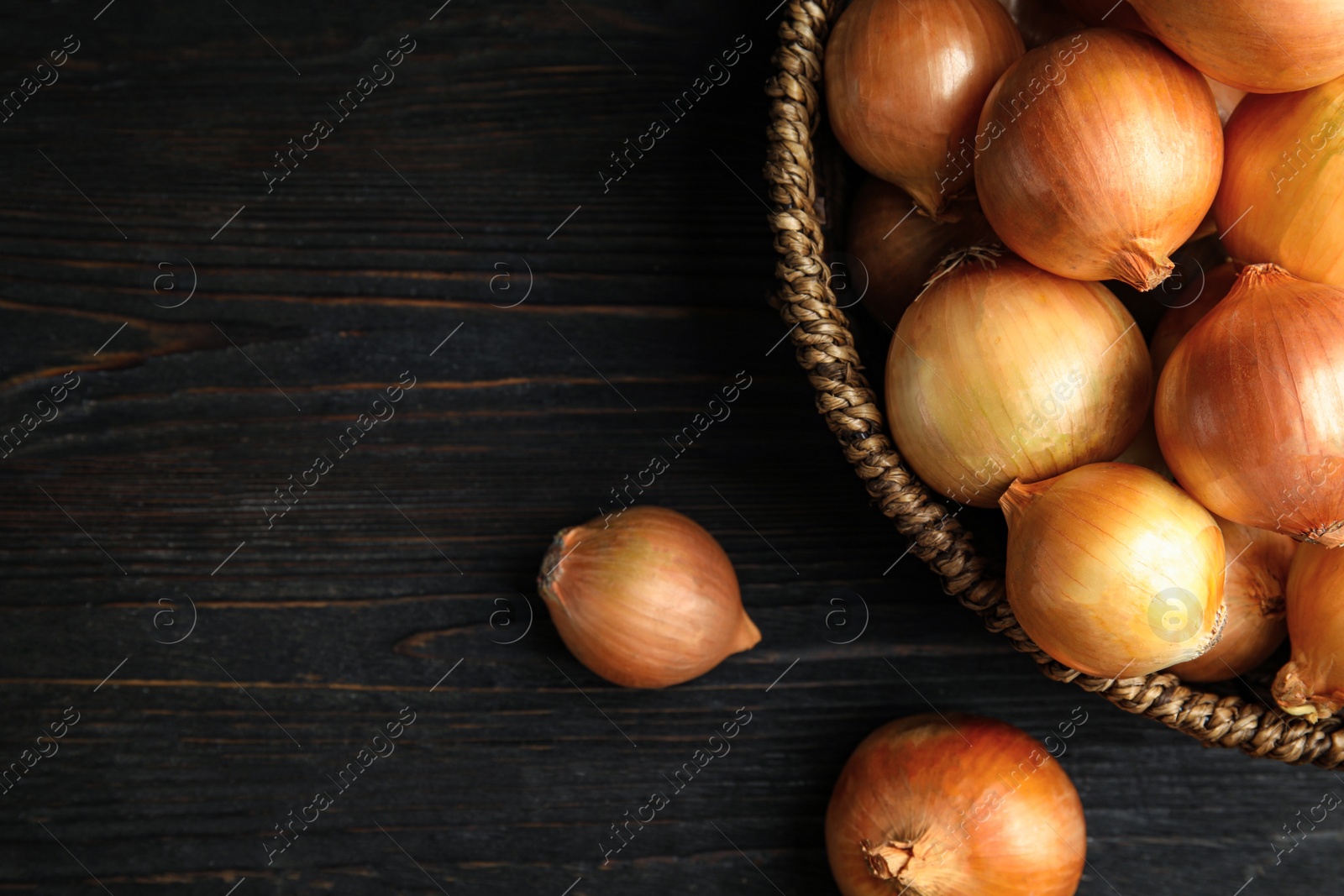 Photo of Wicker basket of ripe onions on black wooden table, flat lay with space for text