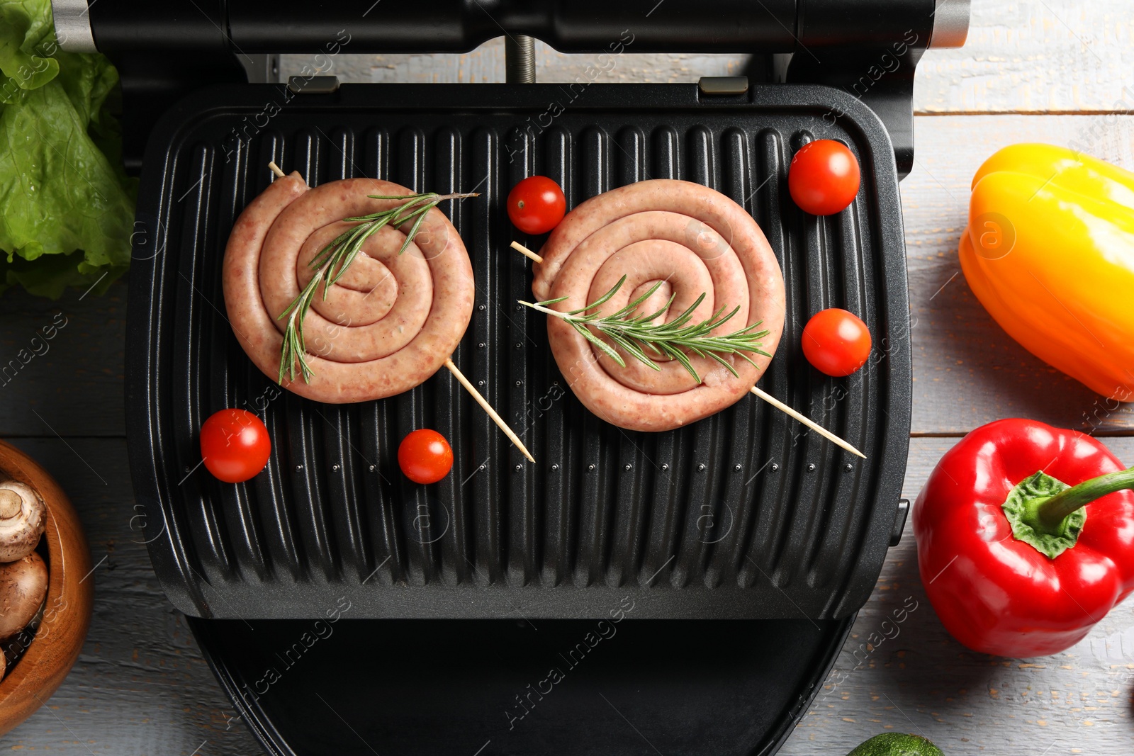 Photo of Electric grill with homemade sausages, rosemary and vegetables on rustic wooden table, flat lay