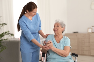 Photo of Nurse giving cup of tea to woman in wheelchair indoors. Assisting senior people