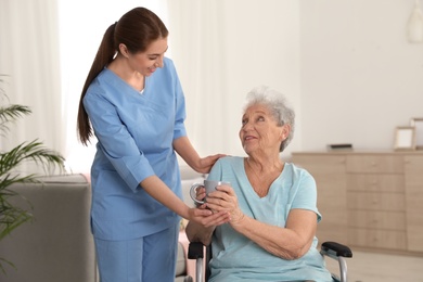 Nurse giving cup of tea to woman in wheelchair indoors. Assisting senior people