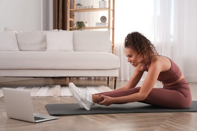 Photo of Beautiful African American woman stretching on yoga mat while watching online class at home