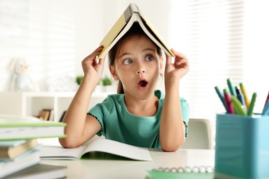 Emotional little girl with book on her head doing homework at table indoors