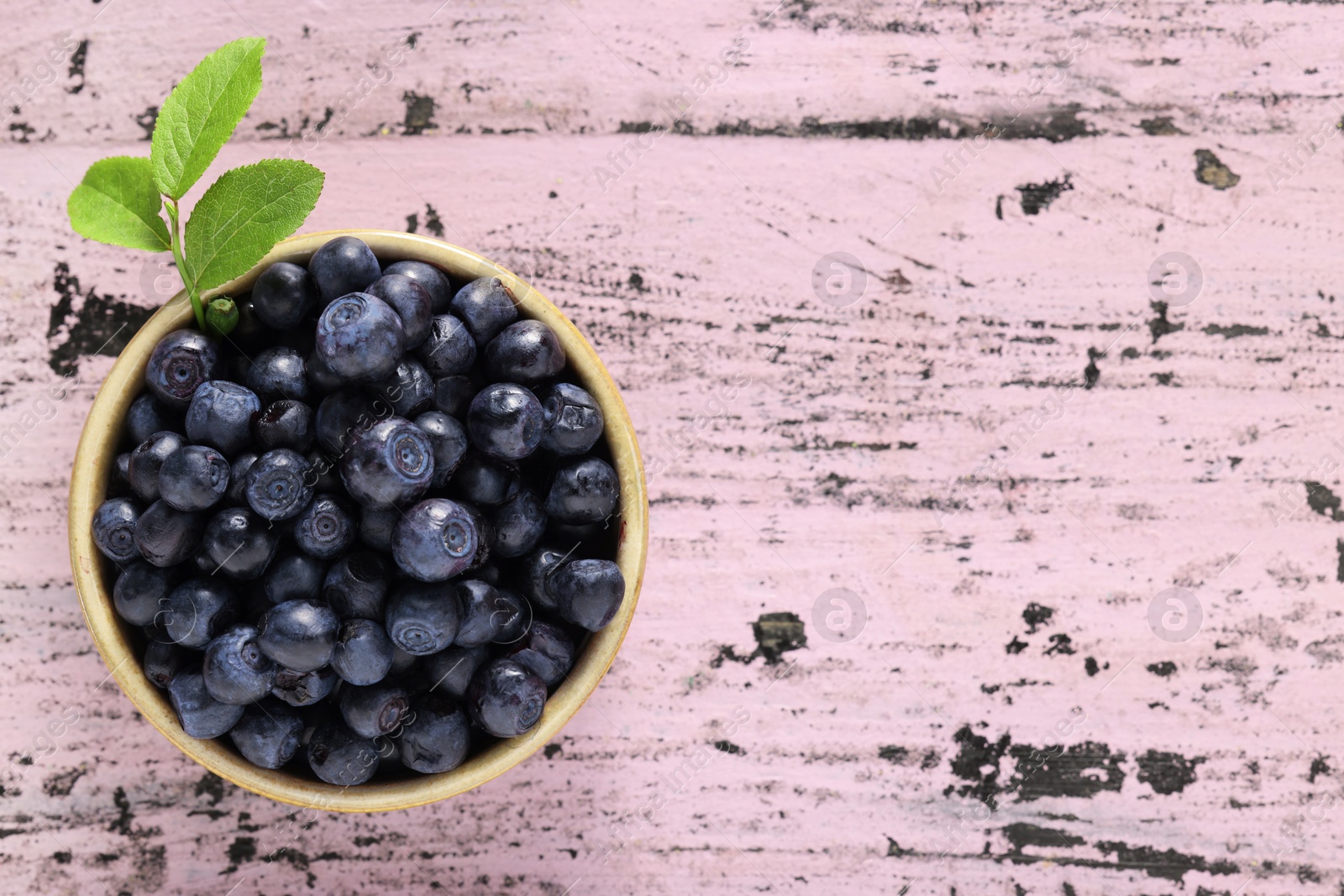 Photo of Tasty fresh bilberries with green leaves in bowl on old pink wooden table, top view. Space for text