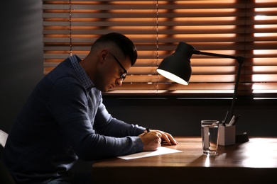 Man writing letter at wooden table in dark room