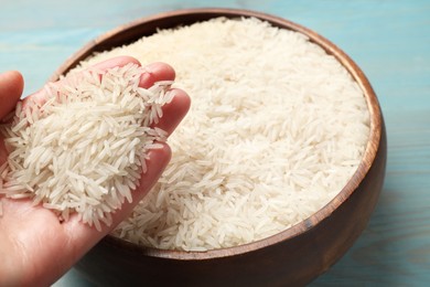 Photo of Woman holding raw basmati rice over bowl at light blue wooden table, closeup