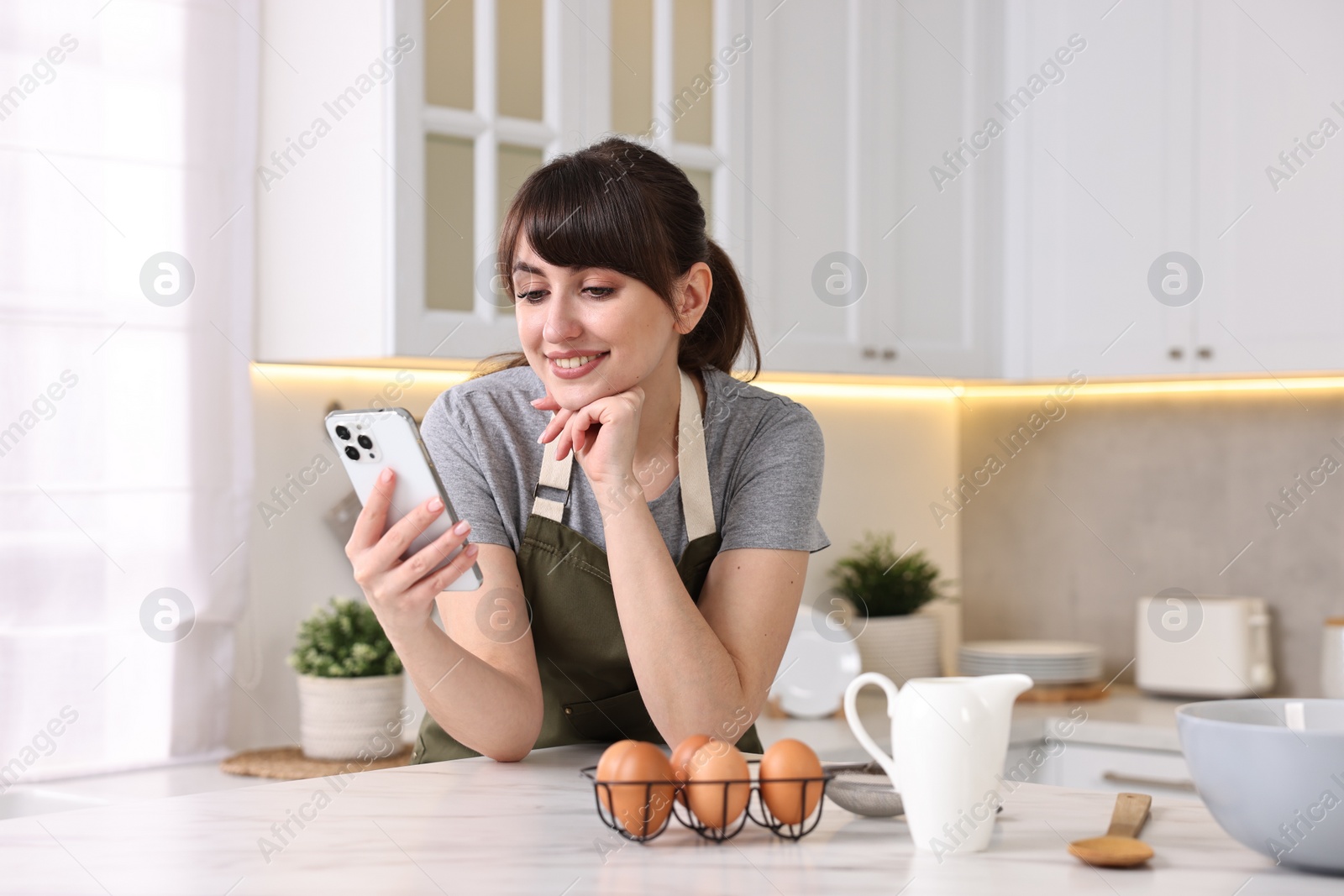 Photo of Happy young housewife using smartphone while cooking at white marble table in kitchen