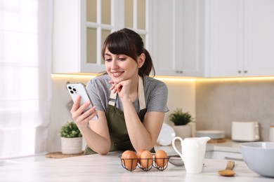 Happy young housewife using smartphone while cooking at white marble table in kitchen