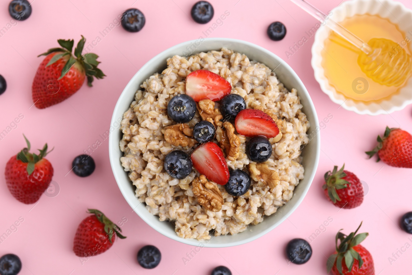 Photo of Tasty oatmeal with strawberries, blueberries and walnuts in bowl surrounded by fresh berries on pink background, flat lay