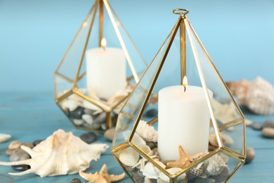 Photo of Stylish glass holders with burning candles, seashells and pebbles on light blue wooden table, closeup