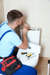 Professional plumber in uniform repairing toilet tank indoors