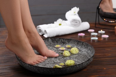 Photo of Woman soaking her feet in bowl with water and flowers on wooden surface, closeup. Pedicure procedure