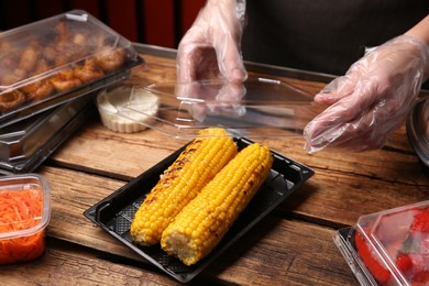 Photo of Waiter in gloves closing container with grilled corn cobs at wooden table, closeup. Food delivery service