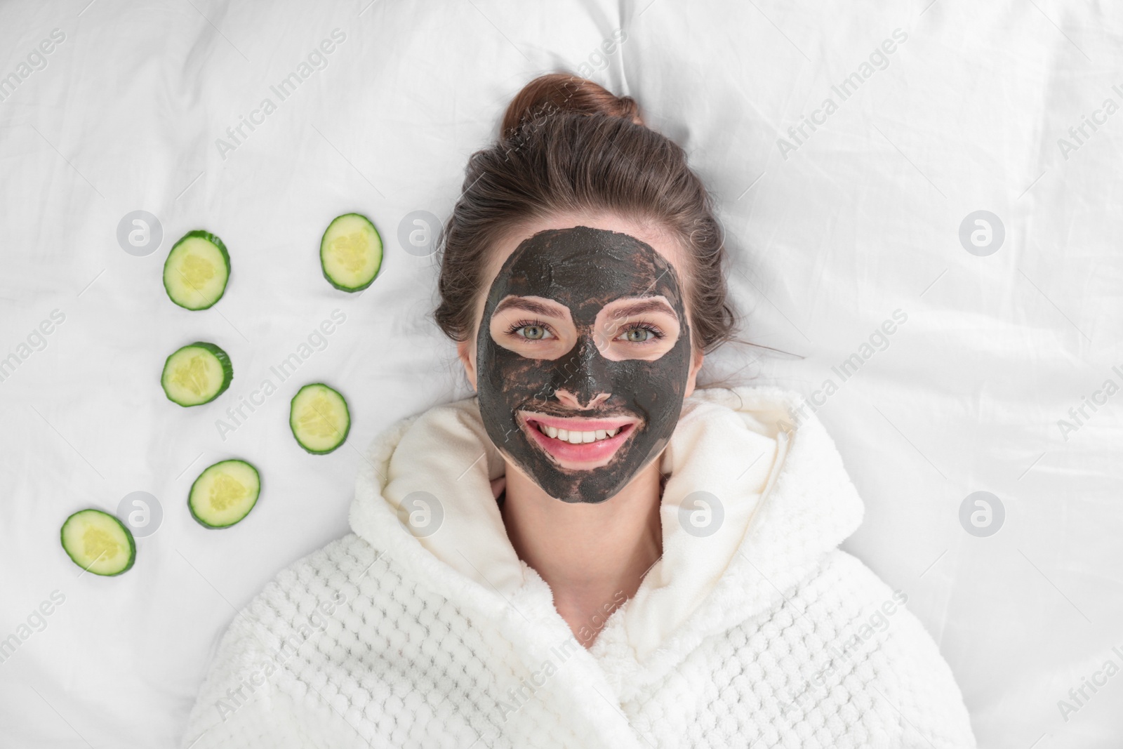 Photo of Young woman with facial mask lying and cucumber slices on bed, top view