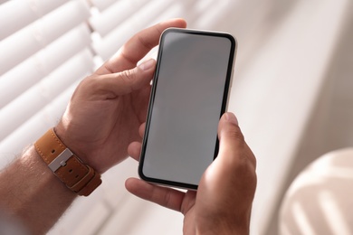 Man holding mobile phone with empty screen near window indoors, closeup