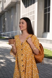 Beautiful young woman with stylish backpack and cup of coffee on city street