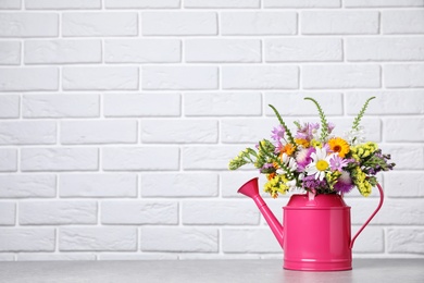 Watering can with beautiful wild flowers on table near brick wall