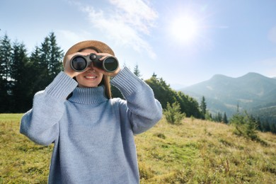 Young woman with binoculars in mountains on sunny day, space for text