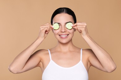 Photo of Beautiful young woman putting slices of cucumber on eyes against beige background