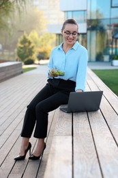 Photo of Smiling businesswoman holding lunch box and working with laptop outdoors
