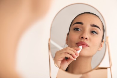 Woman with herpes applying cream on lips in front of mirror against light background