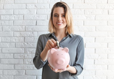 Beautiful woman putting money into piggy bank near brick wall