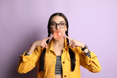 Photo of Fashionable young woman with braids blowing bubblegum on lilac background