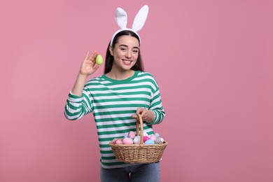 Happy woman in bunny ears headband holding wicker basket of painted Easter eggs on pink background. Space for text