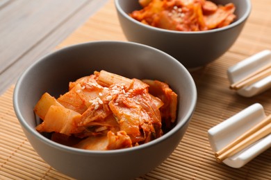 Photo of Bowls of spicy cabbage kimchi and chopsticks on bamboo mat, closeup