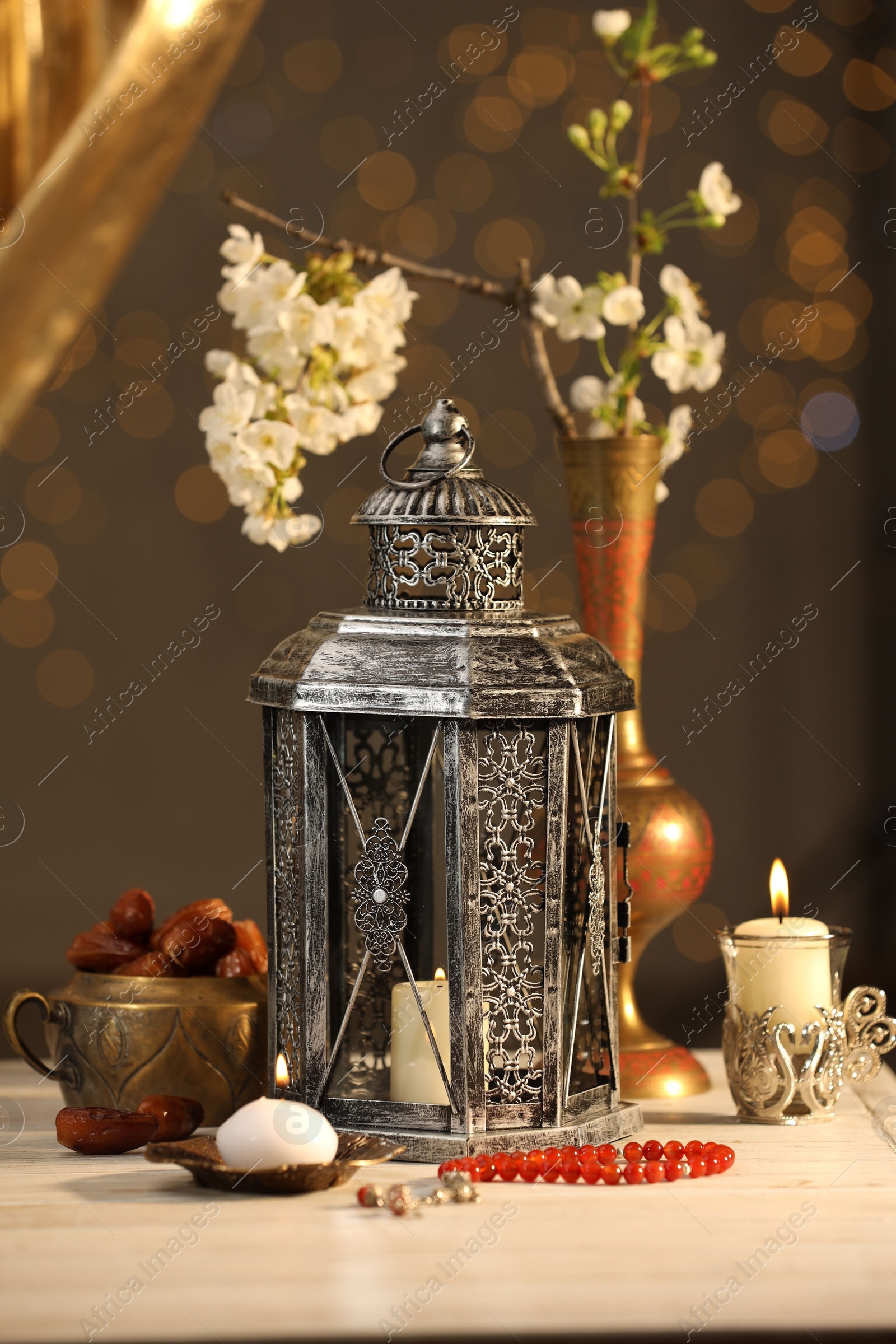 Photo of Arabic lantern, misbaha, candles, dates and flowers on table against blurred lights