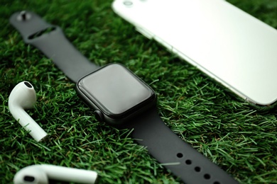 Photo of Stylish smart watch, phone and earphones on green grass, closeup