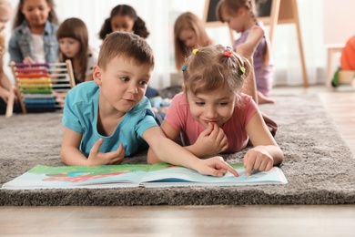 Photo of Cute kids reading book on floor while other children playing together in kindergarten