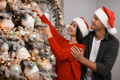 Photo of Happy young couple in Santa hats decorating Christmas tree together at home