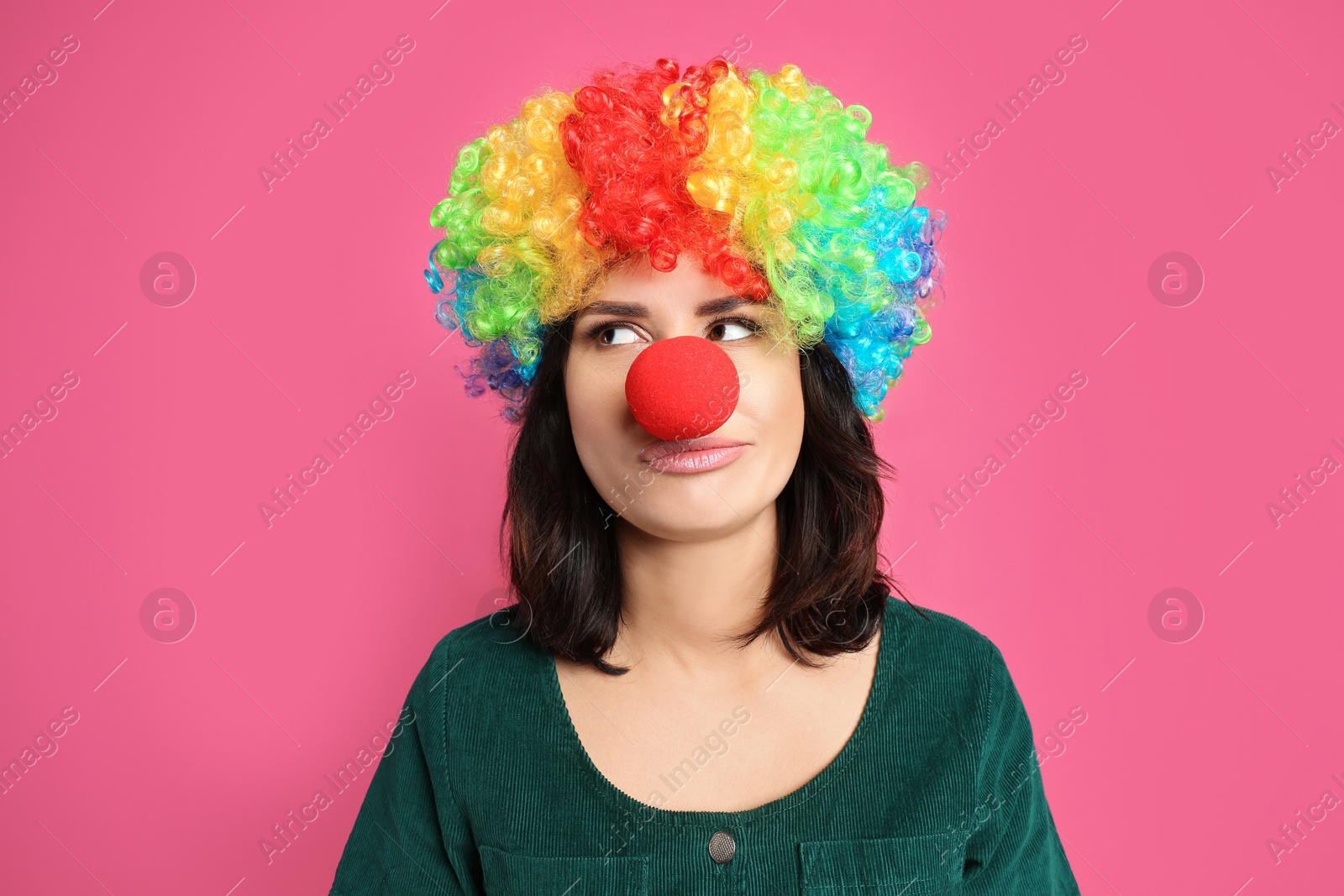 Photo of Emotional woman with rainbow wig and clown nose on pink background. April fool's day