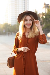 Beautiful young woman in stylish red dress and hat with handbag on city street