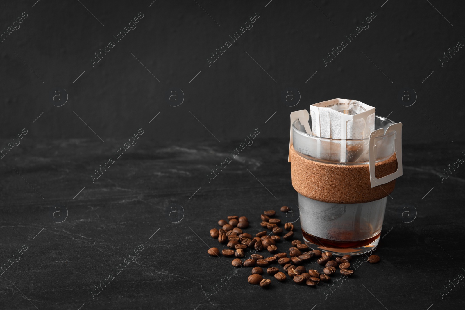 Photo of Glass cup with drip coffee bag and beans on black slate table. Space for text