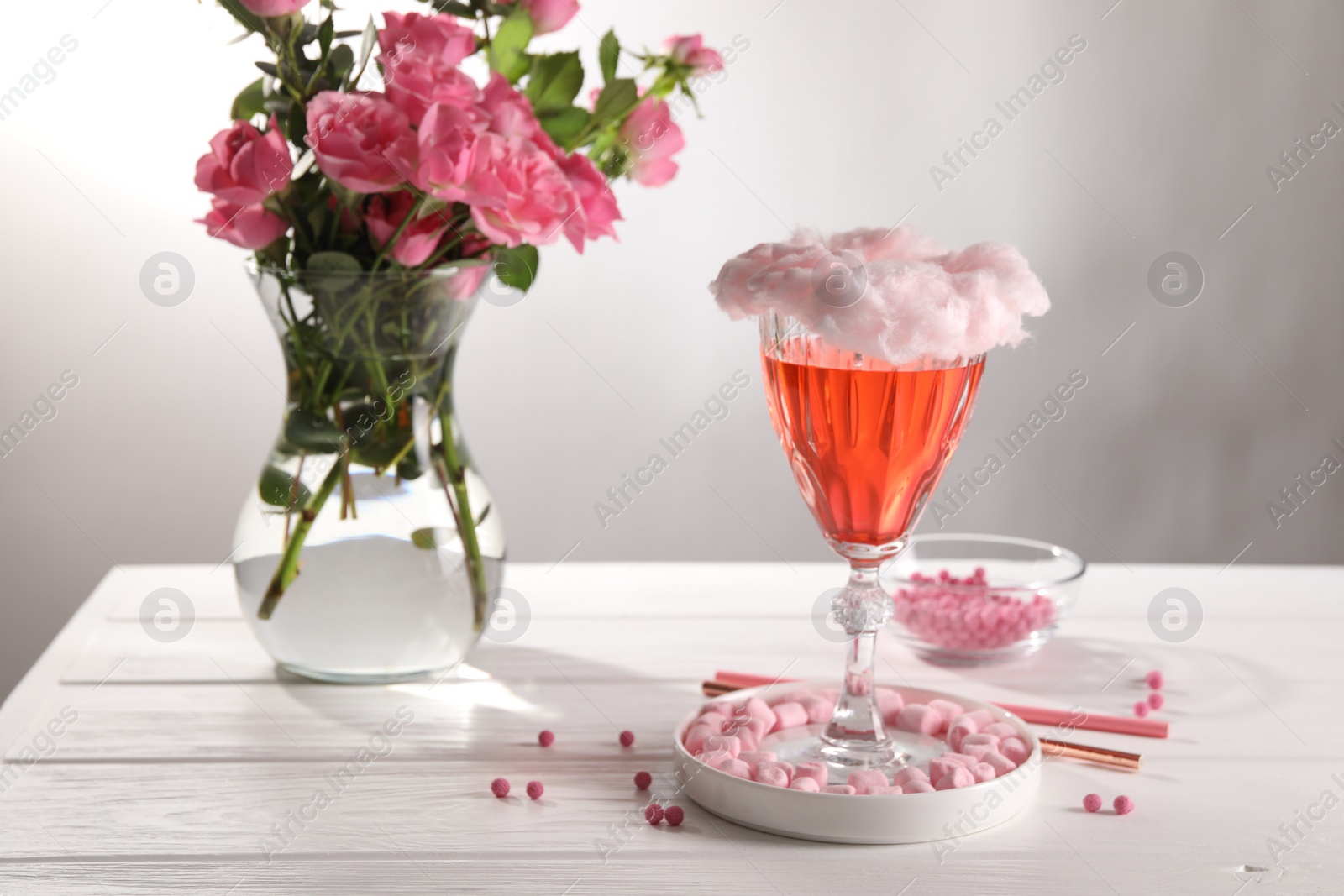 Photo of Cotton candy cocktail in glass, marshmallows, vase with pink roses and straws on white wooden table against gray background