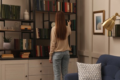 Photo of Young woman choosing book on shelf in home library, back view