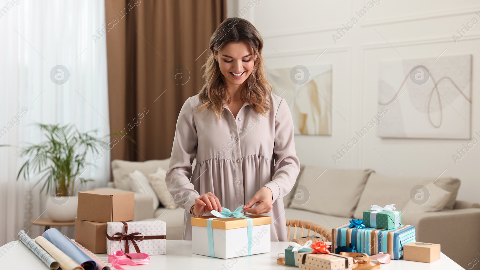 Photo of Beautiful young woman decorating gift box with ribbon at table in living room