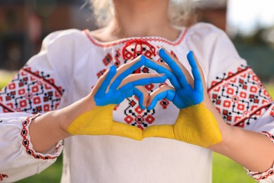 Photo of Little girl making heart with her hands painted in Ukrainian flag colors outdoors, closeup. Love Ukraine concept