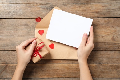 Photo of Woman holding blank greeting card and gift at wooden table, top view. Valentine's day celebration