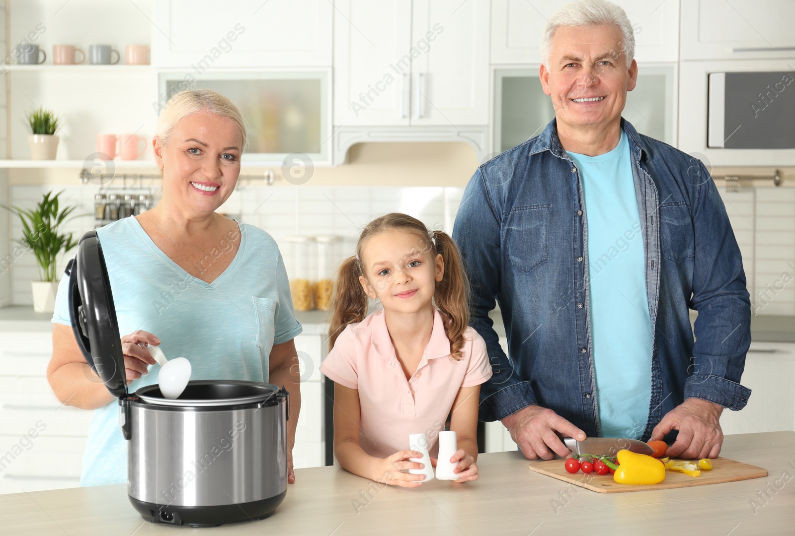 Photo of Mature couple and their granddaughter preparing food with modern multi cooker in kitchen