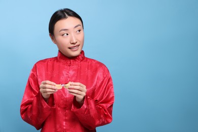 Photo of Asian woman breaking tasty fortune cookie with prediction on light blue background, space for text