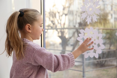 Photo of Little girl decorating window with paper snowflake indoors