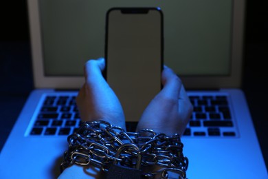 Photo of Woman with chained hands holding smartphone near laptop on dark background, closeup. Internet addiction