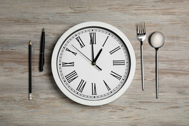 Wall clock, cutlery, pencil and pen on light wooden table, flat lay. Business lunch concept