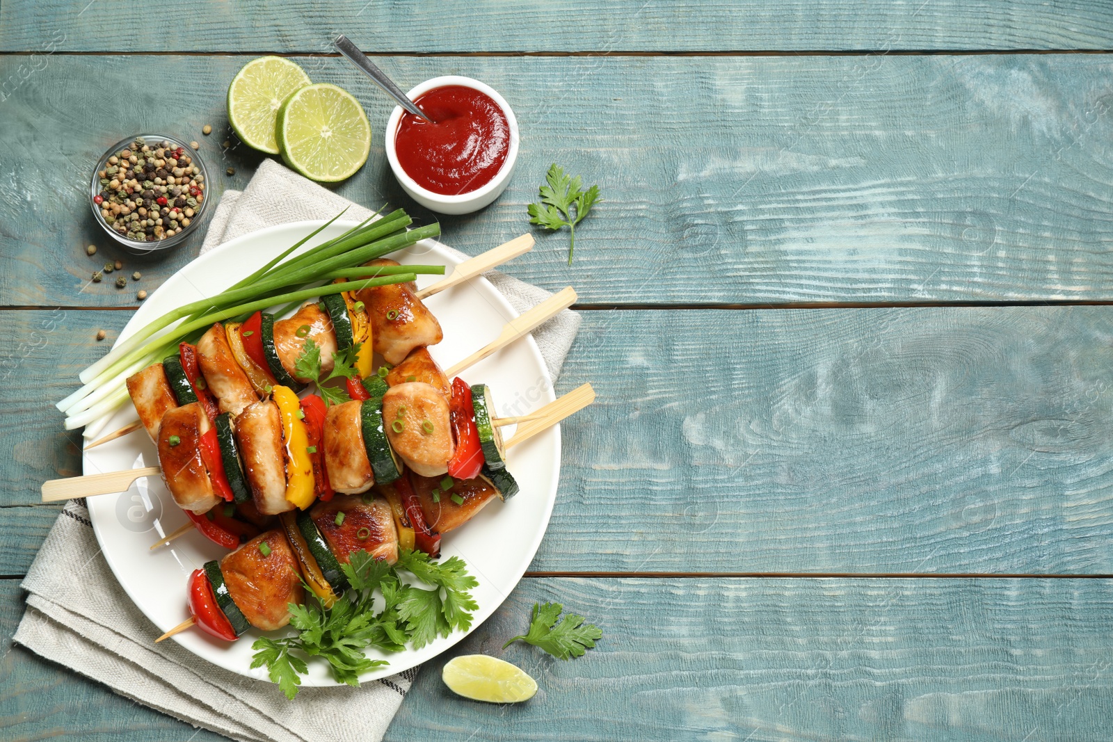 Photo of Delicious chicken shish kebabs with vegetables and ketchup on light blue wooden table, flat lay. Space for text