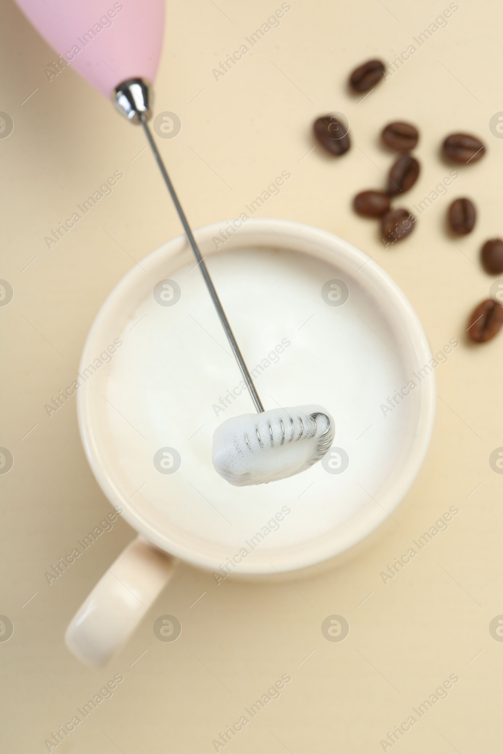 Photo of Mini mixer (milk frother), cup of whipped milk and coffee beans on beige background, flat lay