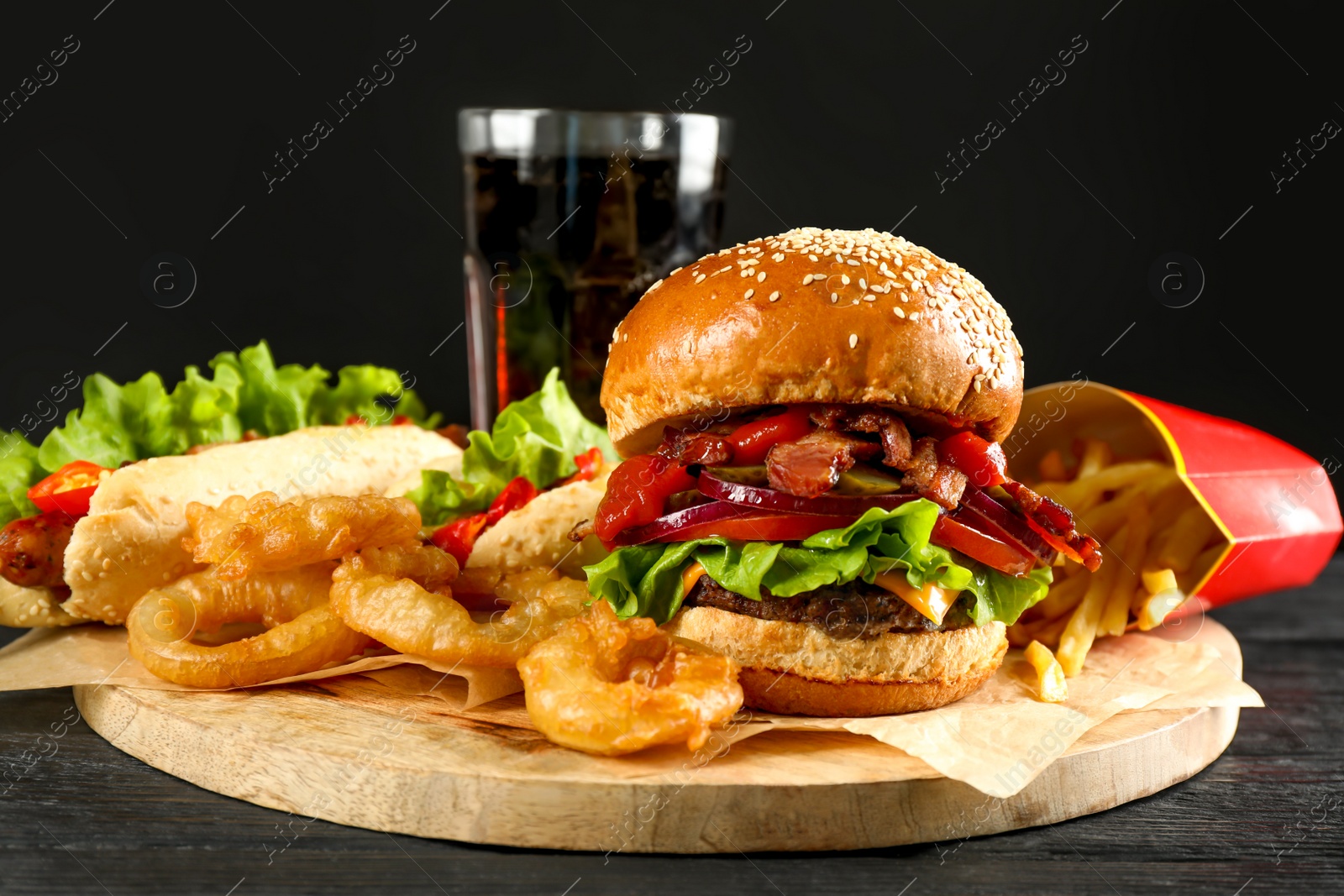 Photo of Tasty burger, French fries, fried onion rings, hot dog and refreshing drink on black wooden table. Fast food