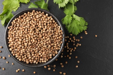 Photo of Dried coriander seeds in bowl and green leaves on dark gray textured table, flat lay